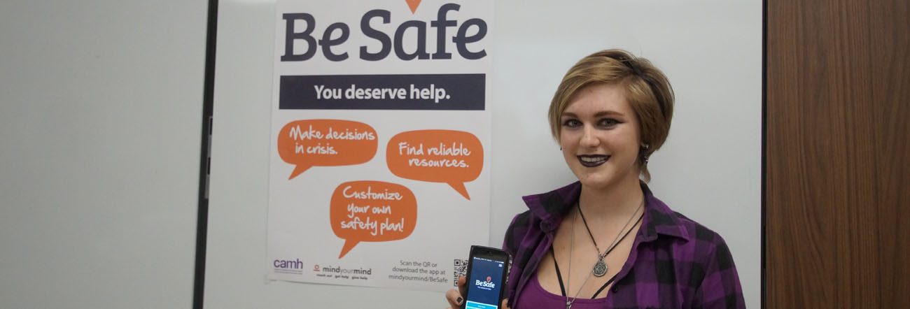 Female student holding cell phone with Be Safe App and Be Safe poster in background