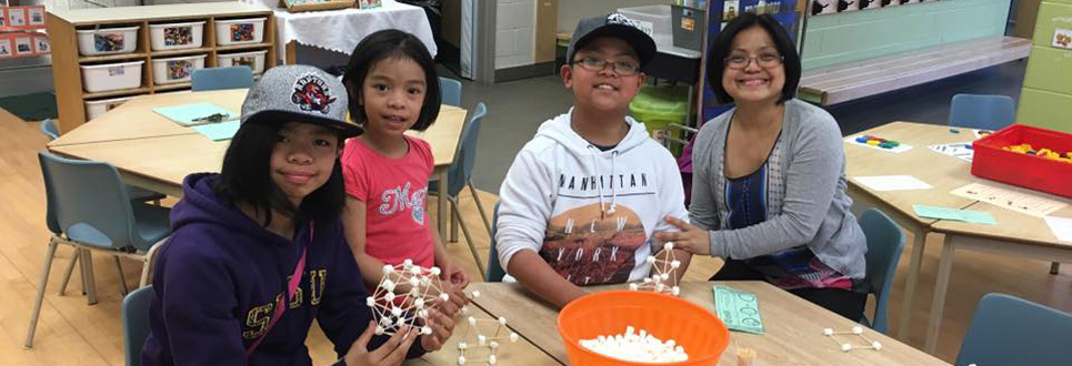 Three students creating geometric shapes with marshmallows and toothpicks with the help of a parent volunteer.